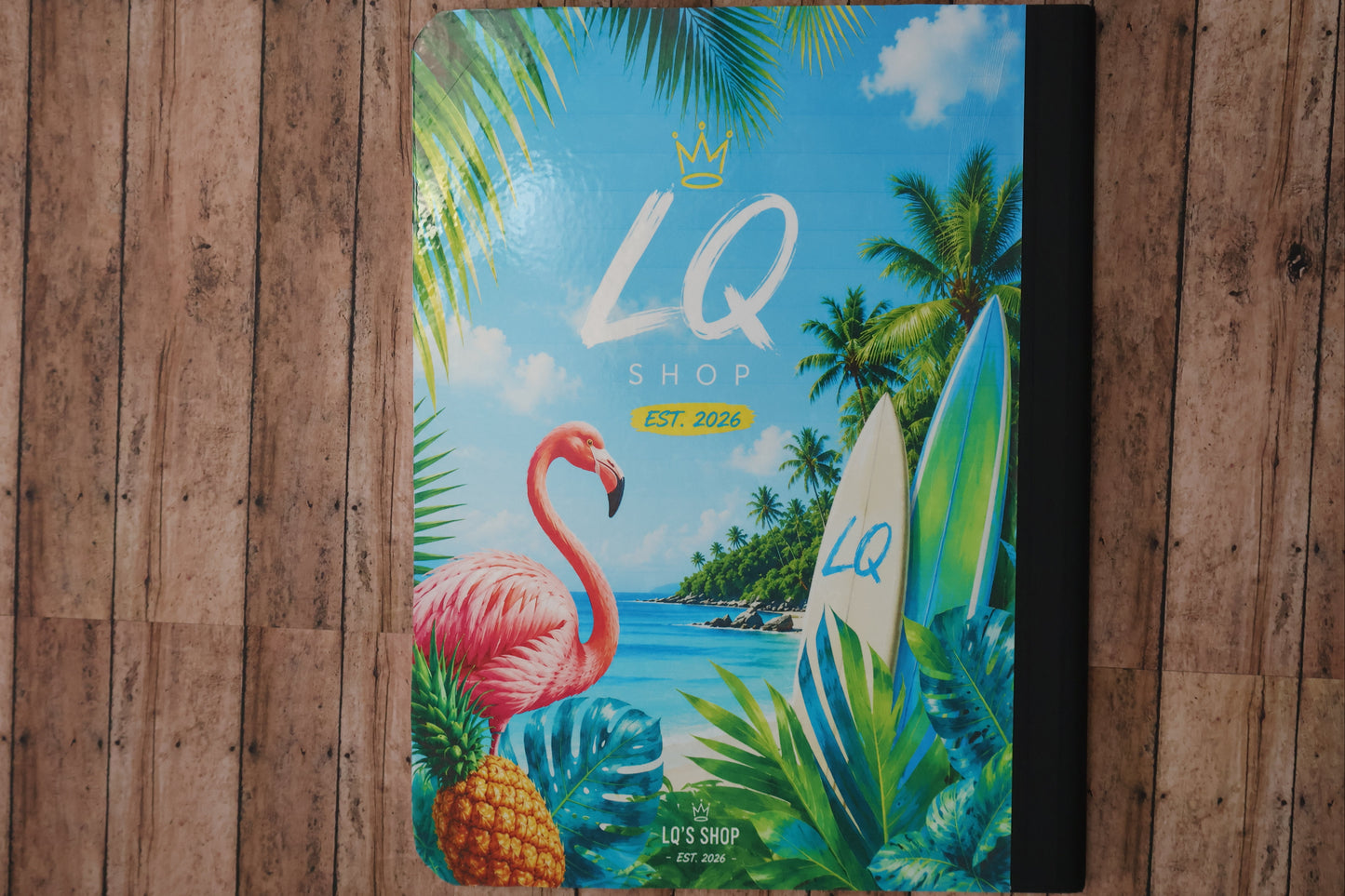 Colorful notebook with tropical design featuring a flamingo, pineapple, and surfboard on a wooden surface.