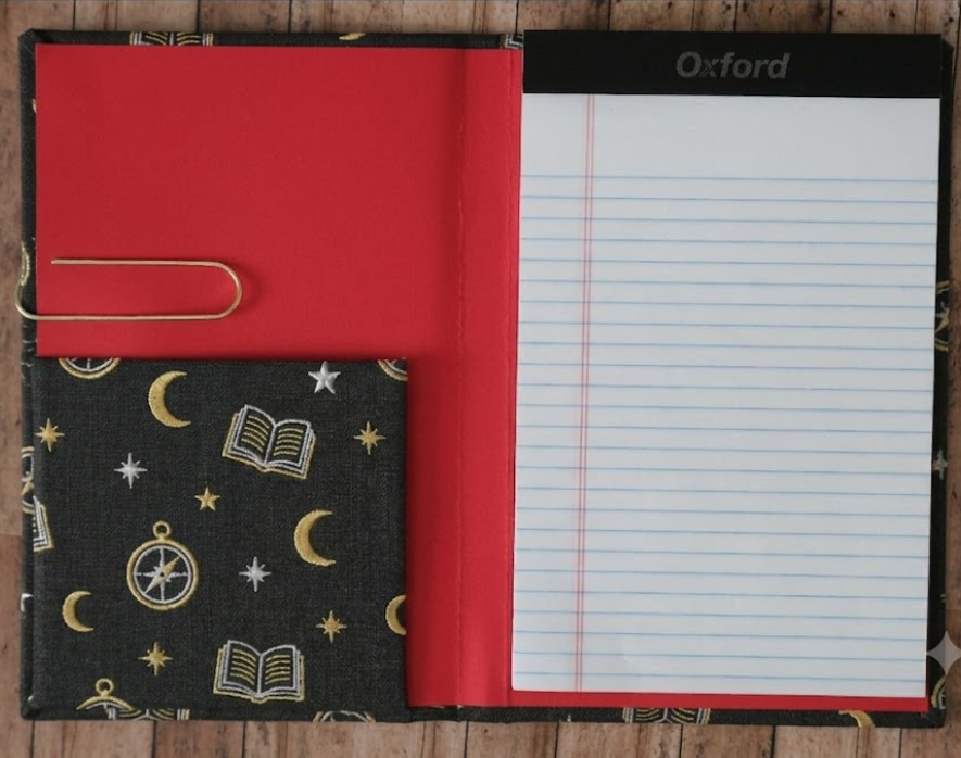 Notebook with red cover and lined pages, black cover with celestial pattern, and gold paper clip on wooden surface.